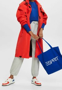 Roter wasserdichter Mantel über einem blauen Strickpullover, grauen Jogginghosen und weißen Sneakers mit roten und blauen Akzenten. Blauer Tote-Bag mit "ESPRIT"-Logo.