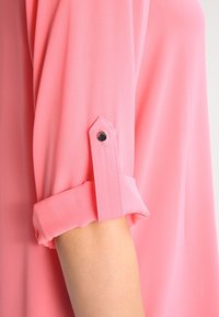 Ljusrosa blus med uppvikt ärm som hålls på plats med en svart knapp, tillverkad av ett mjukt och smidigt tyg med en flödande design.