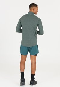 Langarm, grüne Sportbluse mit hohem Kragen, kombiniert mit türkisen Shorts. Strukturierter Stoff, figurbetontes Design und sichtbare Nahtdetails.