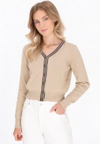 Beige neulejakku, jossa on V-pääntie, ja joka korostuu raitatehosteella reunassa sekä messinkipainikkeilla. Neulottu pinta, jossa on ribbottomit hihansuissa ja helmassa.