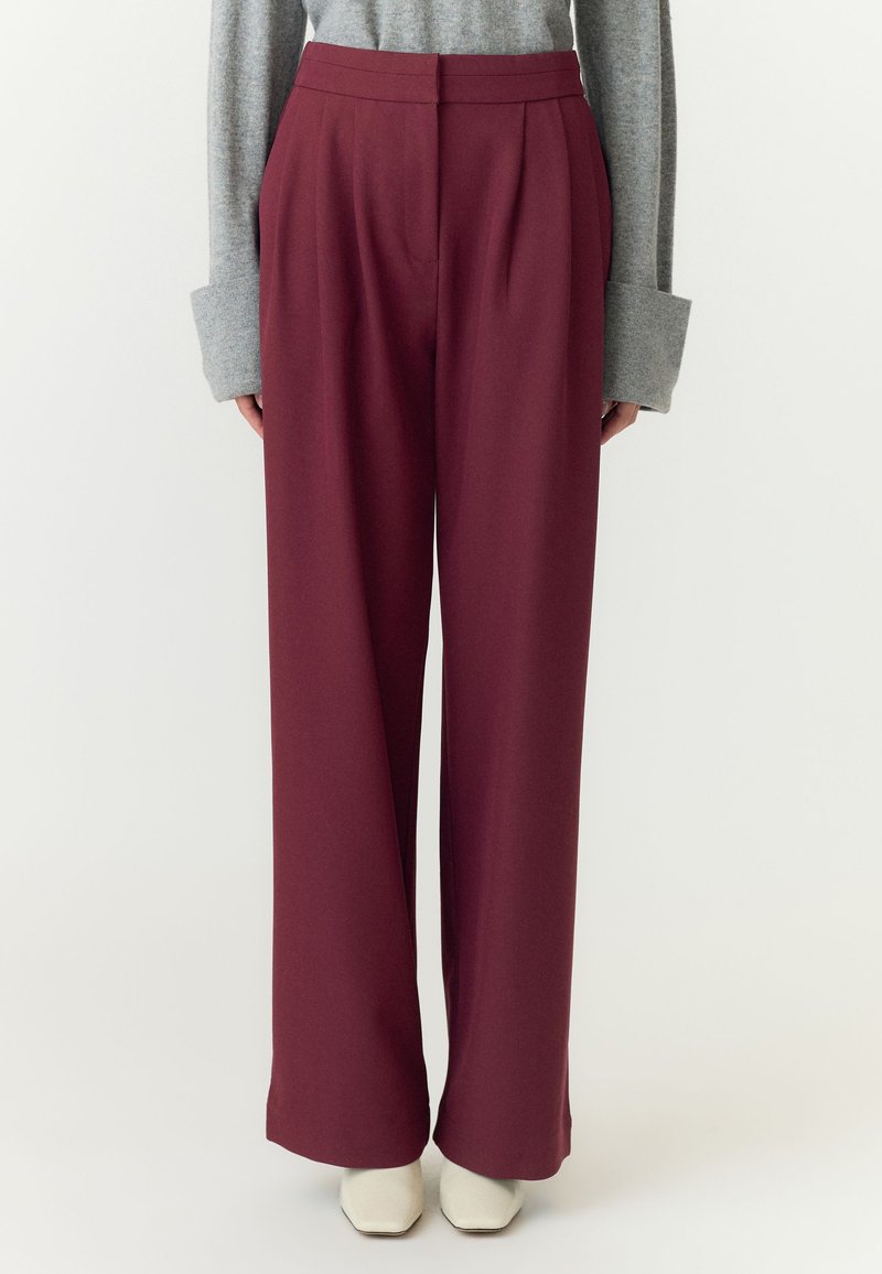 Pantaloni bordeaux a vita alta e gamba larga indossati con una maglia grigia a manica lunga e scarpe bianche con punta quadrata su uno sfondo semplice.