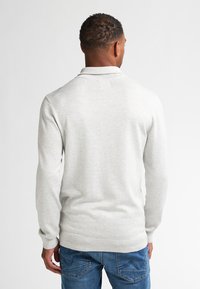 Maglione grigio chiaro a collo alto con polsini ribattuti, caratterizzato da una superficie liscia e un design aderente. Indossato con jeans di denim blu.