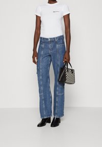 Person som bär en vit åtsittande t-shirt och blå raka jeans, håller en svart-vit mönstrad handväska och svarta stövlar.