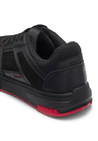 Scarpa sportiva nera con tomaia testurizzata, accenti in mesh e suola in gomma con battistrada rosso a motivo. Logo visibile sul lato.