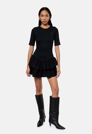 Vrouw draagt een zwarte jurk met korte mouwen en een gerimpelde rok en zwarte kniehoge laarzen met hak, staand tegen een effen lichte achtergrond.