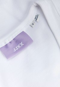 Gros plan sur un tissu blanc côtelé avec une étiquette de marque violette cousue "JJXX" et de petites étiquettes indiquant la taille S et fabriqué au Bangladesh.