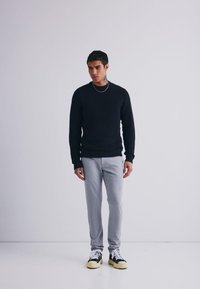Crna teksturirana jumper, svijetlosive uske hlače i crne tenisice s žutim detaljima. Model stoji nasuprot obične pozadine.