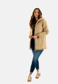 Chaqueta beige con capucha, cierre frontal con cremallera, puños elásticos y bolsillos laterales, llevada sobre una camiseta negra y acompañada de jeans azules y zapatillas beige.