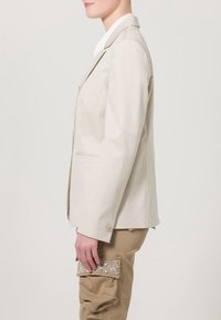 Jasbeżowa marynarka o strukturalnym fasonie, zapinana na jeden guzik, z delikatnymi poduszkami na ramionach. Połączona z beżowymi spodniami cargo i cekinową kopertówką.