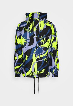 Person trägt eine bunte gemusterte Jacke, schwarze Schneehose und Skischuhe, steht auf einem schneebedeckten Berg und hält Handschuhe in der Hand.