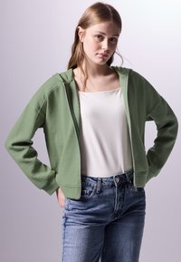 Felpa verde con trama a waffle, dotata di zip, con un taglio rilassato e tasche laterali, indossata sopra una maglietta bianca e abbinata a jeans blu.