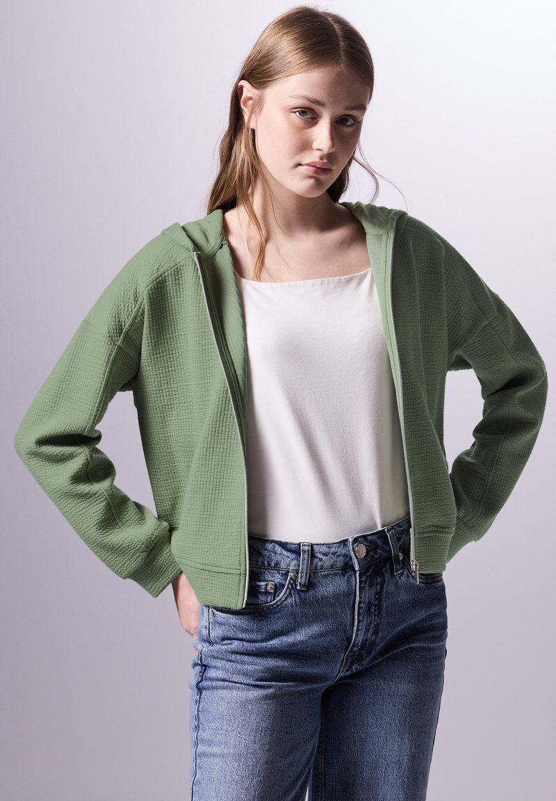 Felpa verde con trama a waffle, dotata di zip, con un taglio rilassato e tasche laterali, indossata sopra una maglietta bianca e abbinata a jeans blu.