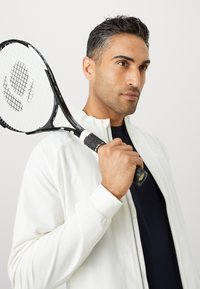 Hvid zip-up jakke over en marineblå skjorte, holder en sort tennisracket med et struktureret håndtag. Baggrunden er ensartet lys grå.