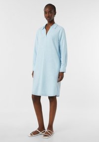 Helles blau gestreiftes Kleid mit langen Ärmeln, Knopfverschluss vorne und seitlichen Schlitzen. Weicher Stoff und lockere Silhouette. Getragen mit weißen Riemchensandalen.