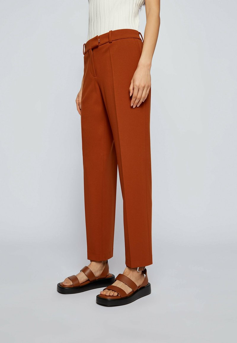 Personne portant un pantalon sur mesure orange brûlé et des sandales en cuir marron assorties, debout sur un fond clair.