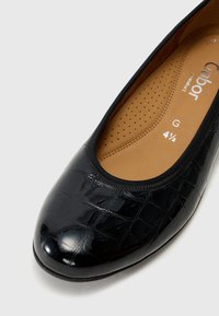 Chaussure plate en cuir verni noir avec une texture crocodile, bout rond, semelle intérieure en cuir beige rembourrée avec des perforations, et bord élastique.