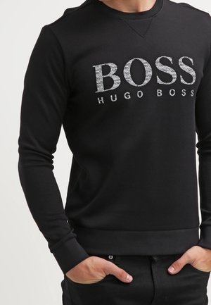 Mies pukeutunut mustaan Hugo Boss -collegepaitaan, jossa suuri valkoinen "BOSS"-teksti ja pienempi "HUGO BOSS"-teksti rinnassa, kädet taskuissa.