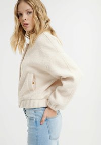 Jasno beżowa krótka kurtka z miękkiego teksturowanego materiału, z kieszeniami po bokach i elastycznymi mankietami, w połączeniu z jasnoniebieskimi dopasowanymi dżinsami.