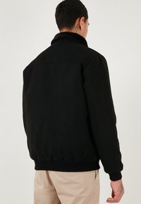 Sort bomberjakke med blød krave, elastiske manchetter og talje, med et enkelt design og glat tekstur. Ingen synlige logoer.