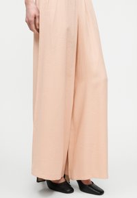 Personne portant un pantalon ample couleur pêche clair et des chaussures noires à talons bas et bouts carrés, debout devant un fond blanc uni.