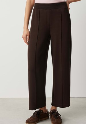 Vrouw draagt donkerbruine wijde broek met voornaad en bruine sneakers, staand tegen een effen lichte achtergrond.