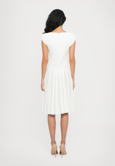 White pleated dress with a fitted bodice and cap sleeves, featuring a back zipper closure. Worn with nude heel sandals.