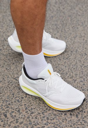 Sneaker atletici bianchi con una tomaia in mesh testurizzato, accenti gialli e neri, e una suola imbottita. Indossati con una calza bianca sopra la caviglia.