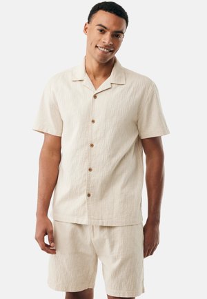 Uomo sorridente, indossa una camicia beige a maniche corte con bottoni e pantaloncini abbinati, in piedi davanti a uno sfondo bianco neutro.