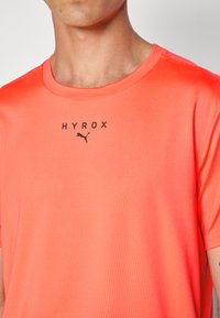 Lys koralfarvet atletisk t-shirt lavet af glat stof. Den har rund hals, korte ærmer og et trykt "HYROX" logo med et lille Puma-emblem.