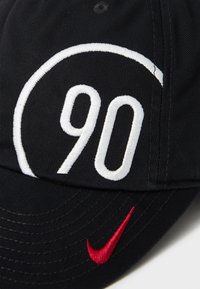 Sort kasket med et hvidt broderet "90" i et cirkulært design og en rød Nike swoosh-logo på siden. Lavet af tekstureret stof.