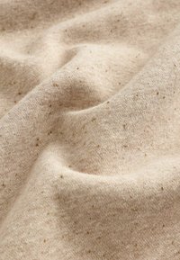 Tissu beige avec de petites taches brunes, montrant des plis doux et une texture tricotée.