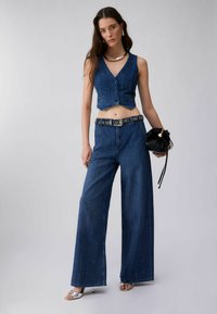 Naine, kes kannab varrukateta lühikest teksavesti ja laia lõikega teksapükse kinnitusdetailidega vööl, hoides musta kurrulist käekotti.