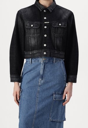Frau trägt eine schwarze, kurze Jeansjacke mit silbernen Knöpfen und einen hoch taillierten blauen Jeansrock mit seitlicher Taschenklappe.