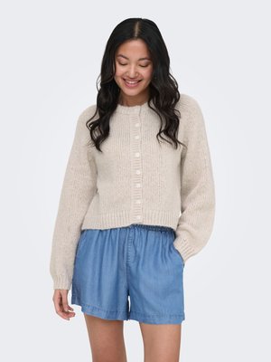Femme portant un cardigan en maille beige boutonné et un short en jean bleu large, debout, une main dans la poche, souriante et regardant vers le bas.