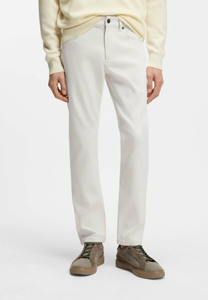 Mężczyzna w jasnobeżowych spodniach, beżowym swetrze i taupe tenisówkach, stojący na jednolitym białym tle.