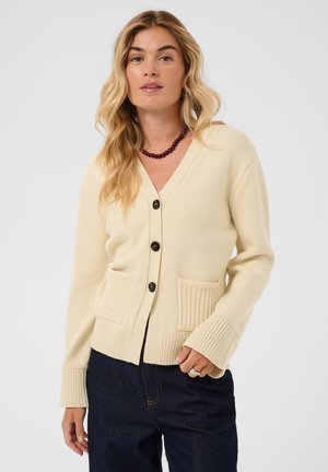 Blonde Frau trägt eine cremefarbene, geknöpfte Strickjacke mit zwei Taschen, dunkelblaue Jeans und eine dunkle Perlenkette vor weißem Hintergrund.