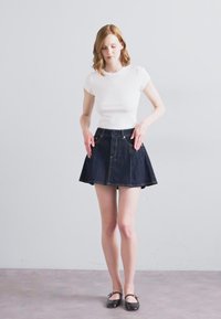 Jupe en denim bleu foncé en forme A avec une taille ajustée et un ourlet évasé. Associée à un haut blanc à manches courtes côtelé et des ballerines noires.
