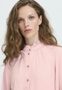Jonge vrouw met golvend bruin haar, gekleed in een lichtroze blouse met knopen en een rimpelcol, die rustig naar de camera kijkt tegen een effen achtergrond.