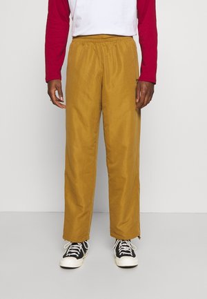 Personne portant un pantalon large jaune moutarde, des baskets noires et blanches, et une chemise blanche avec des manches longues rouges, debout sur un sol gris.