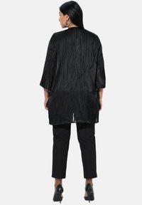 Schwarzer transparenter Kimono mit strukturiertem Muster, dreiviertelärmel und langer Silhouette, kombiniert mit schwarzen cropped Hosen und High Heels.