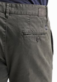 Pantaloni di cotone verde scuro con chiusura a bottone, tasca posteriore e piccolo tag con logo. Presentano cuciture visibili e una texture liscia.