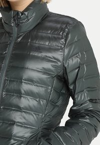 Detailní záběr na osobu, která má na sobě tmavě zelenou prošívanou bundu Adidas s vysokým límcem a předním zipem.