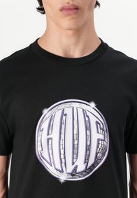 Tricou negru din bumbac, cu un grafic mare în formă de bilă de disc circulară, având cuvântul "HUF" scris cu litere îngroșate, de culoare purpurie. Textură netedă, guler rotund.