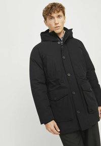La chaqueta parka negra cuenta con una capucha, múltiples bolsillos frontales y cierres de botón. Está hecha de una tela resistente con una textura suave.