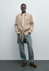 Beige linen shirt over a white t-shirt, light blue jeans, and dark brown suede shoes. Sweater tied around the waist.
