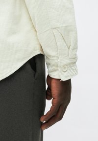 Krém színű bordázott ing gombos mandzsettával, sötétszürke nadrággal párosítva. Az ujjon látható részlet a textúrát és a gombok kialakítását emeli ki.