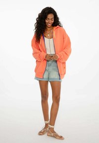 Žiarivo oranžová mikina so zipsom, modrými doplnkami a nastaviteľnými šnúrkami, kombinovaná so svetlým háčkovaným topom a džínsovými šortkami.