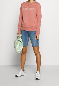 Persona che indossa un felpa rosa di Calvin Klein, pantaloni corti di jeans fino al ginocchio, sneakers bianche e tiene in mano una borsa di stoffa a righe verdi e bianche.