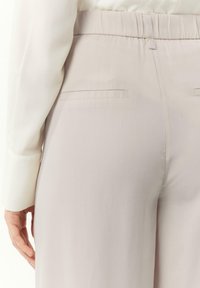 Jasno beżowe spodnie o gładkiej fakturze, z elastycznym pasem i dwiema tylkimi kieszeniami, widoczne w widoku z boku.