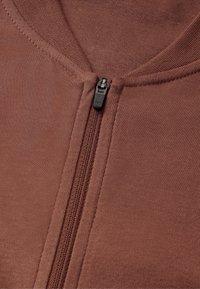 Brun zip-up jacka med en texturerad tyg, med centralt placerad dragkedja och rund krage. Slät yta med tydliga stygn detaljer.